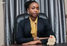 Political Will Is Not Enough. Build the Capability Professional woman seated at a desk in an office, wearing a navy blazer and yellow blouse.