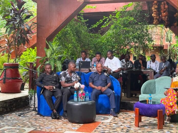 Group of men seated for an outdoor panel discussion under a wooden canopy, microphones and laptops in front, greenery in the background.