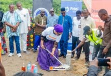 Orange Foundation, Communications Ministry Turn Sod for Transformative Village Project in Bo Woman in a purple headwrap and white shirt breaks ground with a ceremonial shovel at a foundation-laying event, with onlookers and banners in the background.