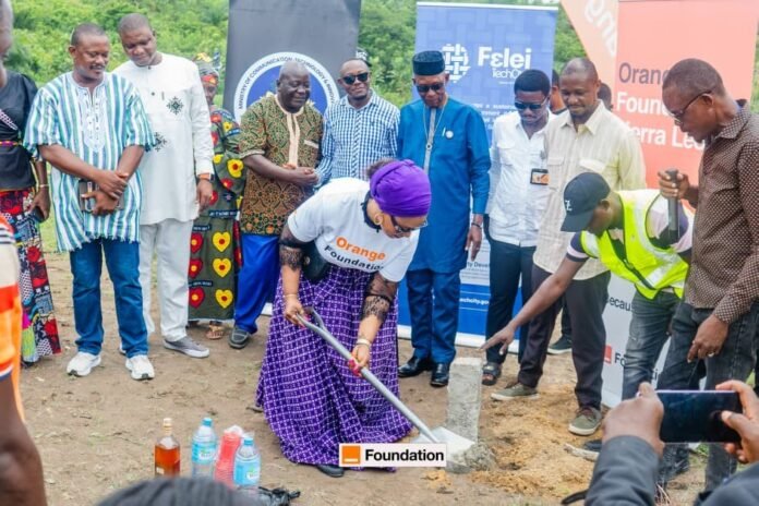 Woman in a purple headwrap and white shirt breaks ground with a ceremonial shovel at a foundation-laying event, with onlookers and banners in the background.
