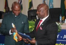 Amb. Fanday Turay Hands Over RMU Leadership Following Two Years of Institutional Transformation Two men in suits exchange a blue folder and a ceremonial item at a formal event, with flags in the background.