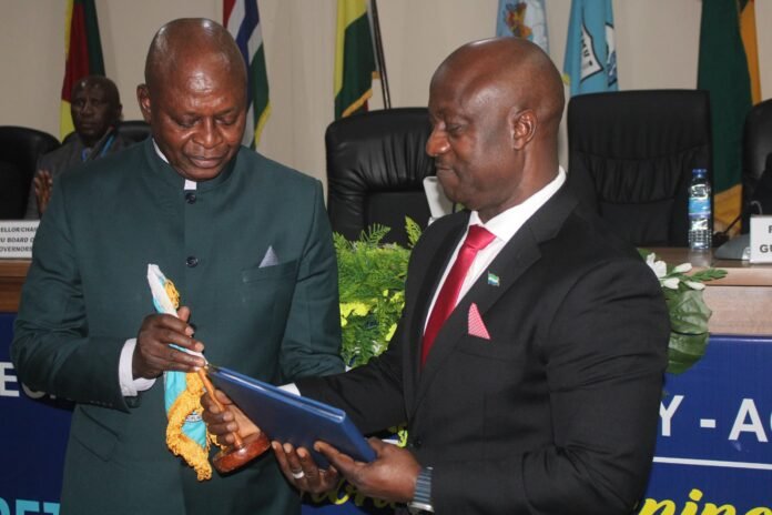 Two men in suits exchange a blue folder and a ceremonial item at a formal event, with flags in the background.