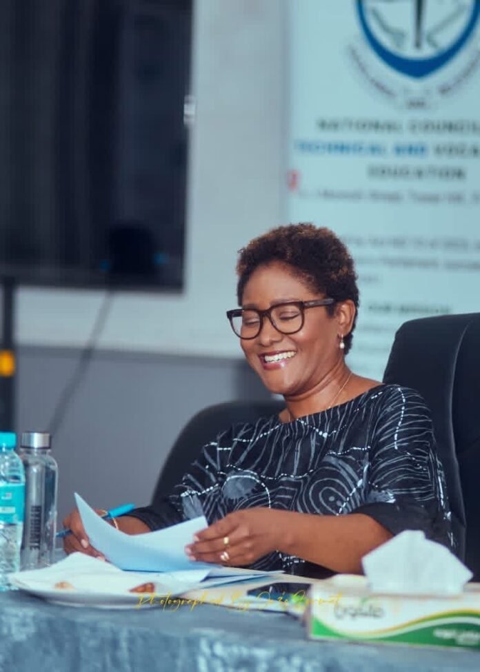 Smiling woman with short curly hair and glasses, seated at a table with papers and a blue pen in a meeting room.