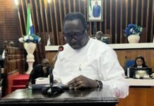 Parliament Ratifies Online Business Registration System Concession to Boost Ease of Doing Business Man in white traditional attire speaks at a wooden podium with a microphone in a formal chamber, with flag and officials in the background.