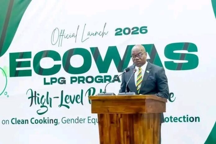Man in a suit delivering a speech at a wooden podium during the ECOWAS LPG Program 2026 official launch, with a banner in green and white behind him. It highlights Clean Cooking and gender equality themes.