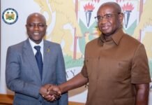 President Bio Engages First Bank Group CEO on Multi-Million-Dollar Infrastructure Financing Initiative Two men shake hands in a formal setting, with a government crest visible in the background on the wall behind them and a round seal on the left edge.