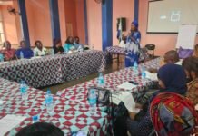 50/50 Group Empowers Women to Champion Climate Action and Food Security Group of people seated around patterned tables in a meeting room, a woman in a blue dress speaks at the front with a microphone.