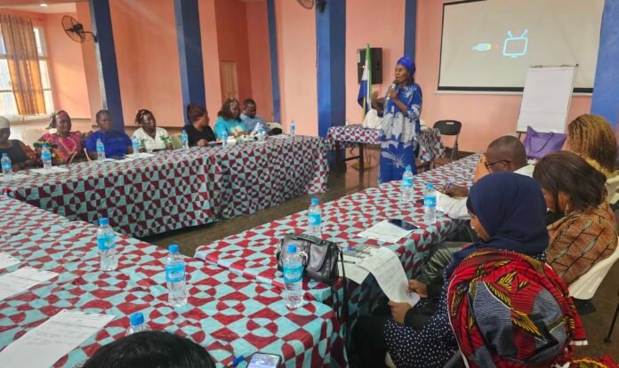 Group of people seated around patterned tables in a meeting room, a woman in a blue dress speaks at the front with a microphone.