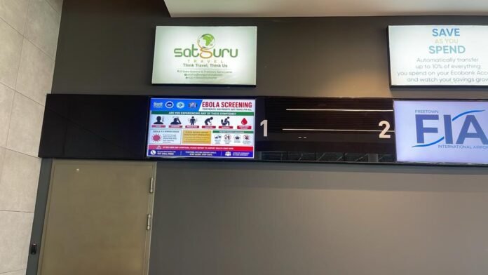 Airport interior with multiple backlit signs: SatSuru Travel logo, an Ebola screening poster, and numbered gates 1 and 2 on a dark wall panelage beside a doorway.