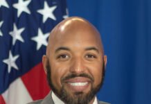 U.S. Embassy Dismisses Visa Closure Rumours, Confirms Normal Operations in Sierra Leone Smiling man in a gray suit and red tie, with an American flag blurred in the background, portrait-style image.