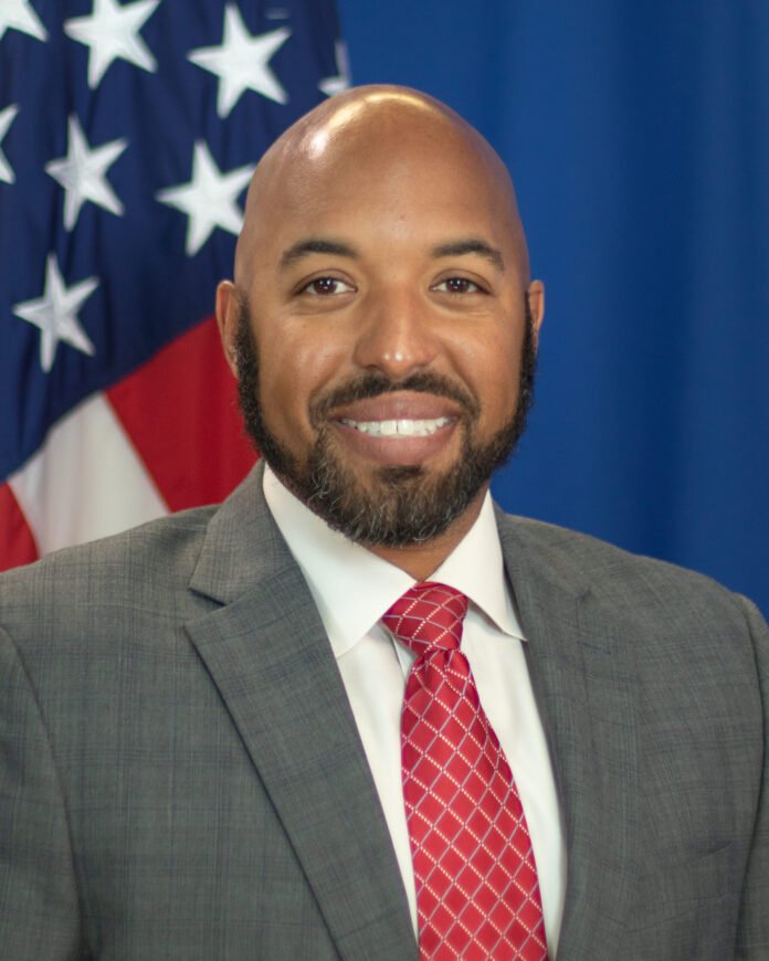 Smiling man in a gray suit and red tie, with an American flag blurred in the background, portrait-style image.