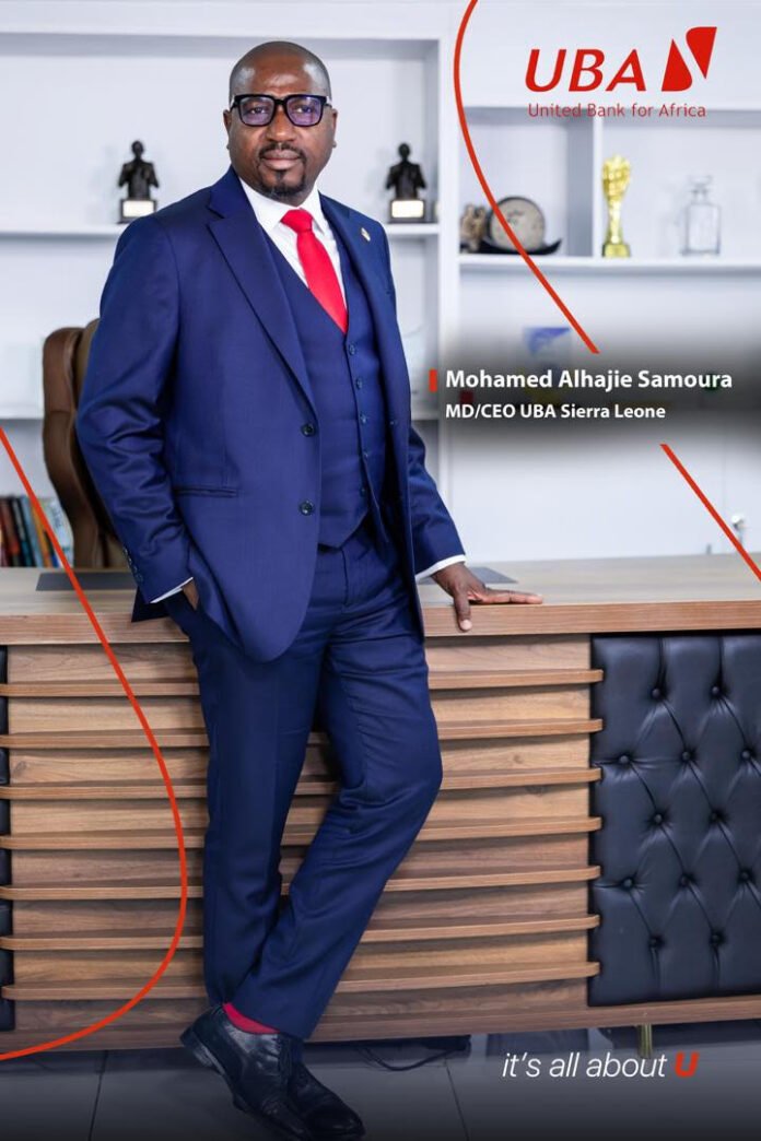 Executive portrait: man in a blue suit and red tie standing beside a wooden desk in a UBA Sierra Leone office with branding in the background, awards on the shelf behind him