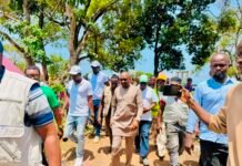 Vice President Inspects Ongoing Additional Classroom Construction Projects in Port Loko District A group of people walking outdoors under trees, some wearing safety vests and hard hats, as a person records with a smartphone.