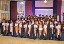 As Over 40 Participants Earn Certificates… Zion Praise Tabernacle & Victorious School of Empowerment Host Maiden Skills Graduation Ceremony Group of graduates in black gowns with orange stoles holding certificates at a church-hall commencement, a cross on the wall behind them.