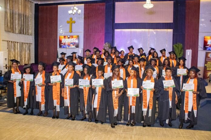 Group of graduates in black gowns with orange stoles holding certificates at a church-hall commencement, a cross on the wall behind them.