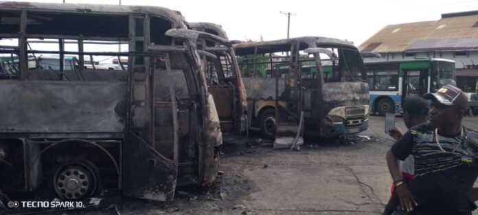 Charred, burnt buses in a yard with doors open and ash on the ground; a man in a cap takes a selfie on the right side of the frame.