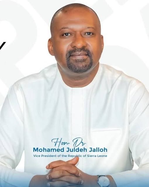 Portrait of Hon. Dr. Mohamed Juldeh Jalloh in a white traditional outfit, hands folded, Vice President of Sierra Leone (text on shirt)