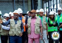 Lungi Conference Centre Nears Completion as Sierra Leone Prepares for ECOWAS Summit Group of officials and workers in hard hats and safety vests touring a construction site, with a man in a pink suit at center addressing the group.