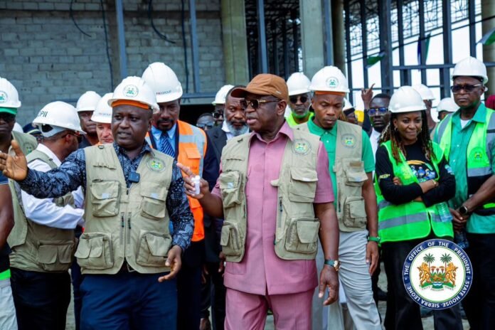 Group of officials and workers in hard hats and safety vests touring a construction site, with a man in a pink suit at center addressing the group.