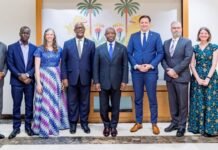 Mercy Ships Honours President Bio for Three Years of Healthcare Collaboration Nine professionals in formal attire pose for a group photo in a reception area, with a palm mural behind them on the wall.