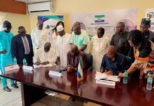 Sierra Leone, So Afri Loge Sign Landmark Housing Agreement for Public Servants Officials sign documents at a formal meeting, with delegates standing around a long table and banners in the background.
