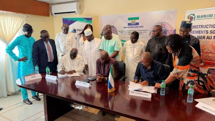 Officials sign documents at a formal meeting, with delegates standing around a long table and banners in the background.