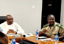 Internal Affairs Ministry Strengthens Security Coordination Through Regular Inter-Agency Meeting Two men sit at a conference table; one in white traditional attire and the other in a tan military uniform holding a microphone.
