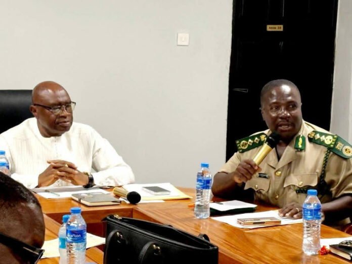 Two men sit at a conference table; one in white traditional attire and the other in a tan military uniform holding a microphone.