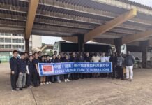 27th Chinese Medical Team Conducts Free Health Clinic for Sierra Leone Government Workers Group of people in dark shirts posing under a concrete canopy, holding a blue banner that reads 'China Medical Team to Sierra Leone' with buses in the background.