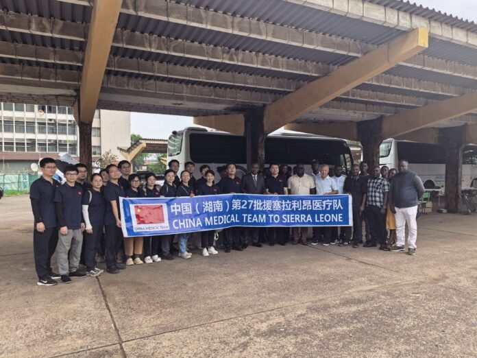 Group of people in dark shirts posing under a concrete canopy, holding a blue banner that reads 'China Medical Team to Sierra Leone' with buses in the background.