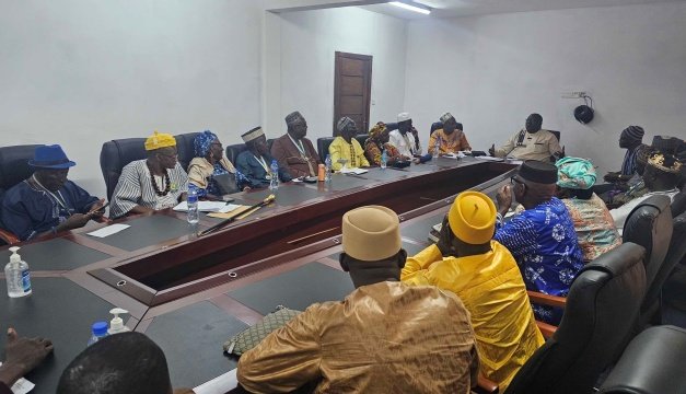 Group of people in traditional attire seated around a long conference table in a meeting room, engaged in discussion with papers and water bottles on the table.