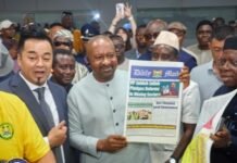 Vice President Reopens Modernized Government Printing Department, Says Public Institutions Can Rise Again Man in a light gray traditional suit holds up a copy of the Daily Mail newspaper while a crowd gathers and a photographer records the moment.