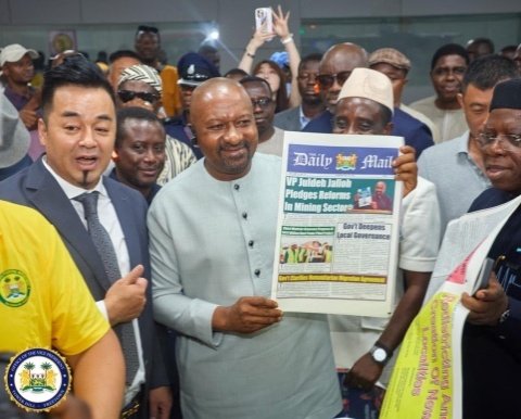 Man in a light gray traditional suit holds up a copy of the Daily Mail newspaper while a crowd gathers and a photographer records the moment.