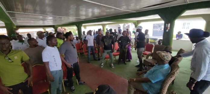 A group of people standing and mingling in a covered outdoor hall with green pillars and red chairs.