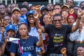 Diverse crowd at a rally, smiling and posing for the camera with raised fists in casual clothing, showing solidarity.