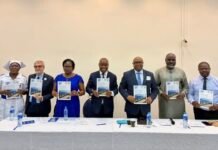 PCMH Records 22% Reduction in Maternal Deaths, Targets Near Zero Cases by 2026 Seven people in formal attire pose behind a long table, each holding a blue-and-white booklet at a ceremonial event.