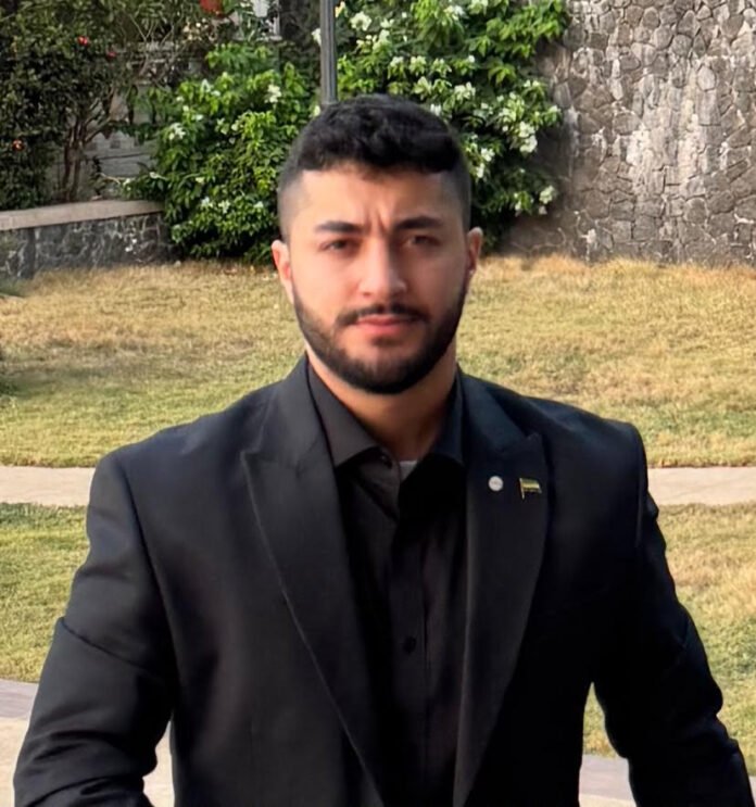 Portrait of a man with short dark hair and a beard, wearing a black suit, standing outdoors near a stone wall and greenery.