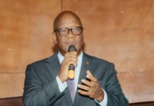 Sierra Leone Records Major Health Gains as 300 Days of Activism Campaign Delivers Results Man in a dark suit and glasses speaks into a handheld microphone at a conference panel; nameplate reads Dr. Austin Demby on the desk in front of him.