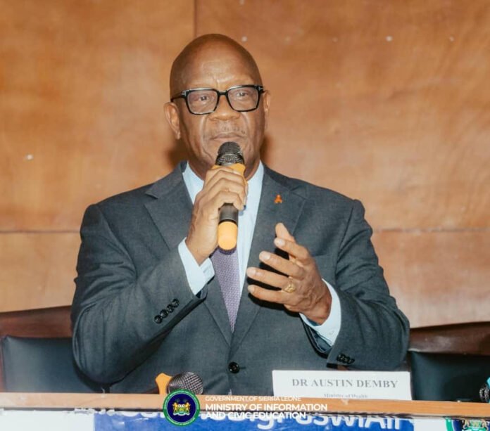 Minister of Health, Dr. Austin Demby Man in a dark suit and glasses speaks into a handheld microphone at a conference panel; nameplate reads Dr. Austin Demby on the desk in front of him.