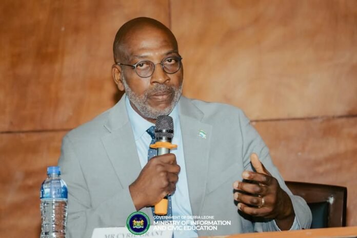 Minister of Energy, Cyril Arnold Grant Man in a light-gray suit speaks into a handheld microphone at a panel, with a water bottle nearby and a Sierra Leone emblem on the table.