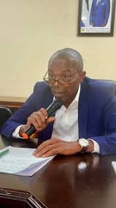 Minister of Lands, Housing and Country Planning, Dr. Turad Senesie Man in a blue suit speaks into a handheld microphone at a desk with papers in front.