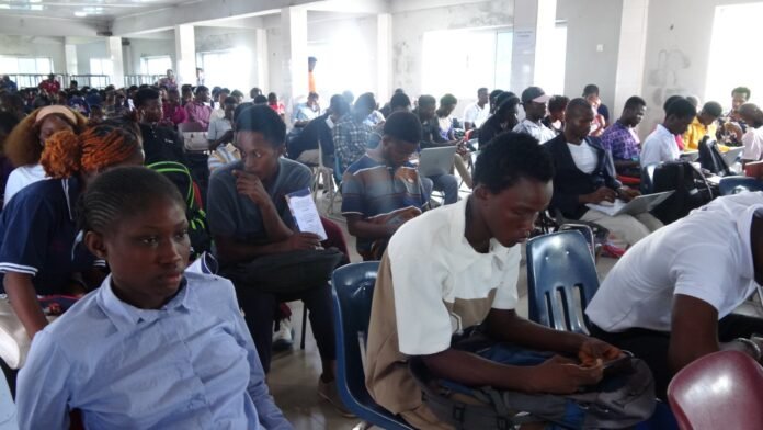 Large hall filled with students seated in rows, many with backpacks and laptops, waiting for an event.