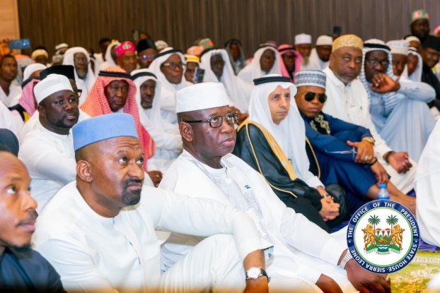 A large crowd of men in white traditional dress seated in a conference hall; central figure wears glasses and a white cap.