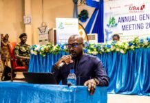 UBA CEO Calls for Development Journalism and Sustainable Media at SLAJ Annual Meeting Man speaker wearing dark blue outfit, holding a microphone at a blue-draped podium during a conference with a laptop and water bottle nearby.