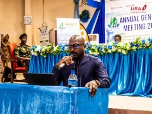 Managing Director and Chief Executive Officer of UBA Sierra Leone and President of the Sierra Leone Bankers Association, Mohamed Alhajie Samoura | The Calabash Newspaper Man speaker wearing dark blue outfit, holding a microphone at a blue-draped podium during a conference with a laptop and water bottle nearby.