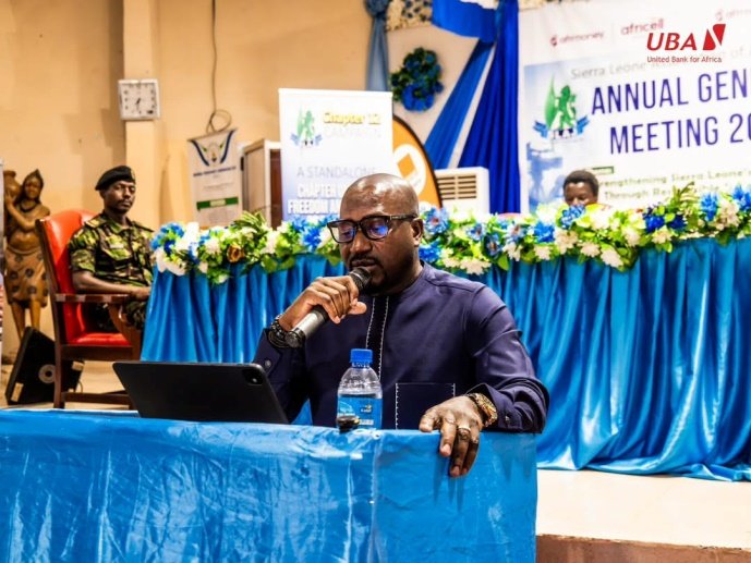 Managing Director and Chief Executive Officer of UBA Sierra Leone and President of the Sierra Leone Bankers Association, Mohamed Alhajie Samoura Man speaker wearing dark blue outfit, holding a microphone at a blue-draped podium during a conference with a laptop and water bottle nearby.