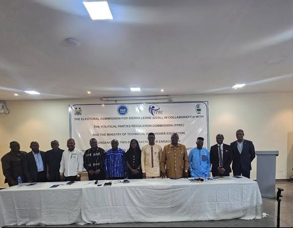 Group of officials standing behind a long table in a conference room, with an Electoral Commission banner behind them.