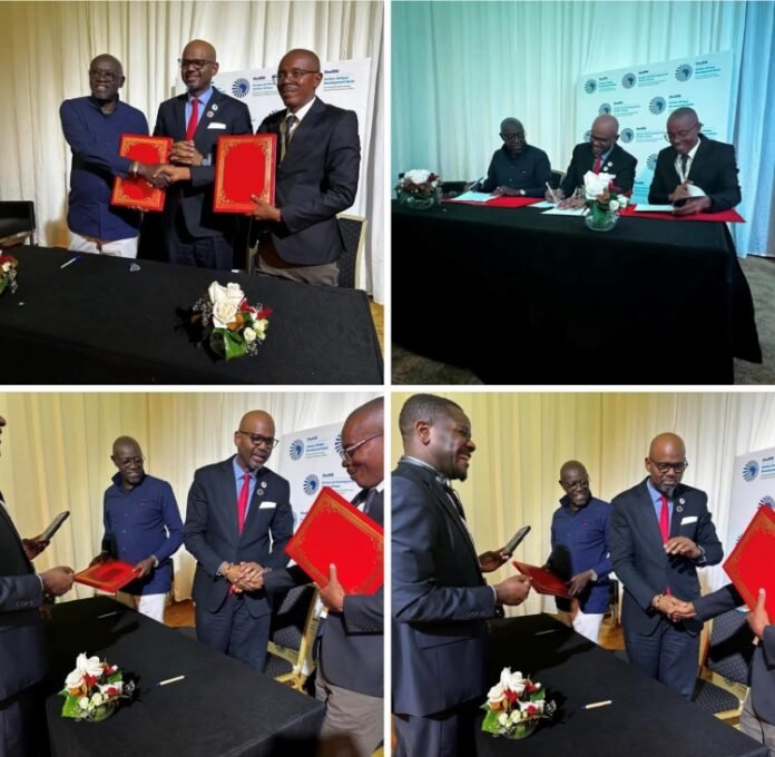 Three men in suits exchange red folders at a formal signing table in a ceremony setting.