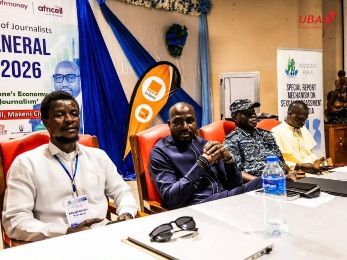 Four men sit at a conference panel table, with banners and a water bottle visible in the background.