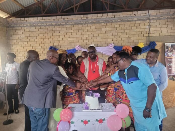 A group of people place their hands together over a cake in a celebratory gathering with colorful balloons in front of a brick wall.